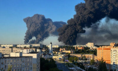 City skyline with enormous black smoke plumes rising from a distant fire across the horizon.