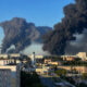 City skyline with enormous black smoke plumes rising from a distant fire across the horizon.