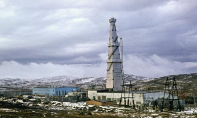 Tall lattice communications tower rising beside blue industrial buildings in a snow-dusted, barren landscape.