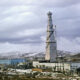 Tall lattice communications tower rising beside blue industrial buildings in a snow-dusted, barren landscape.