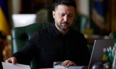 Man in a black shirt reviews a document at a desk with a laptop nearby in a formal office setting.