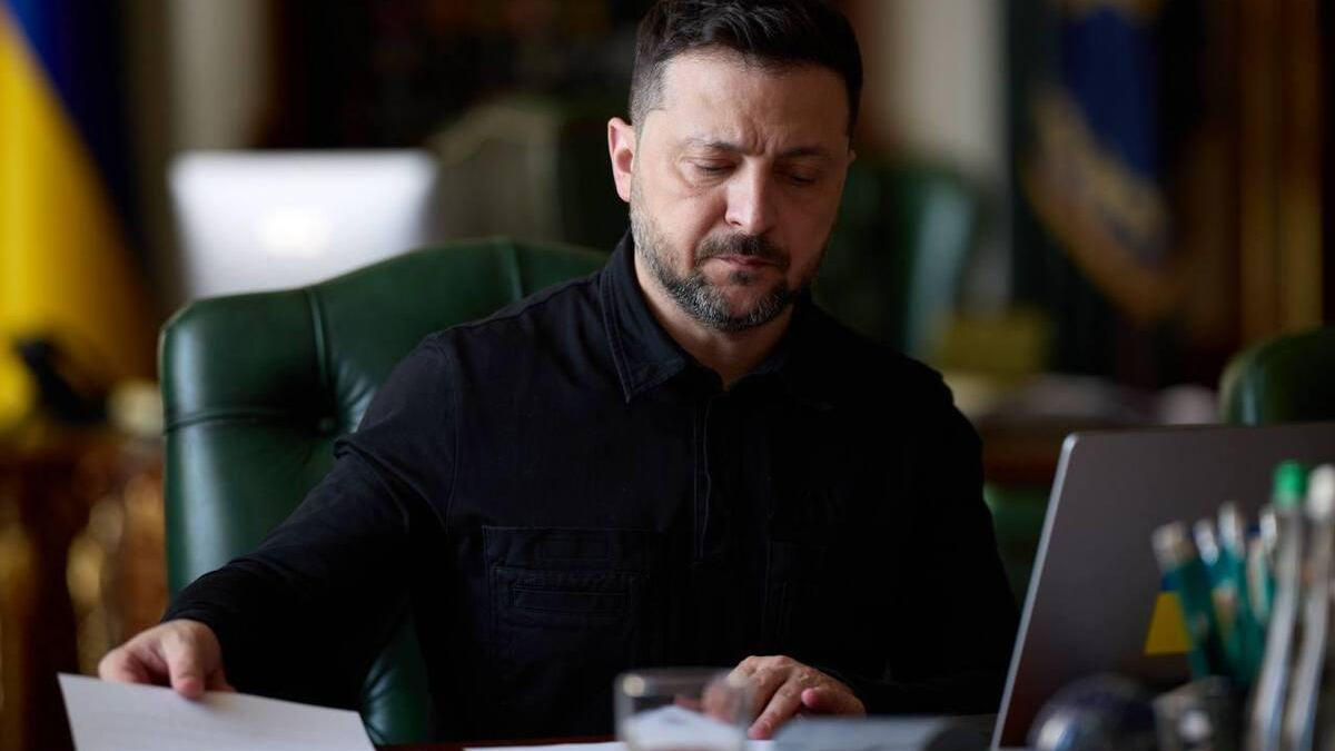 Man in a black shirt reviews a document at a desk with a laptop nearby in a formal office setting.