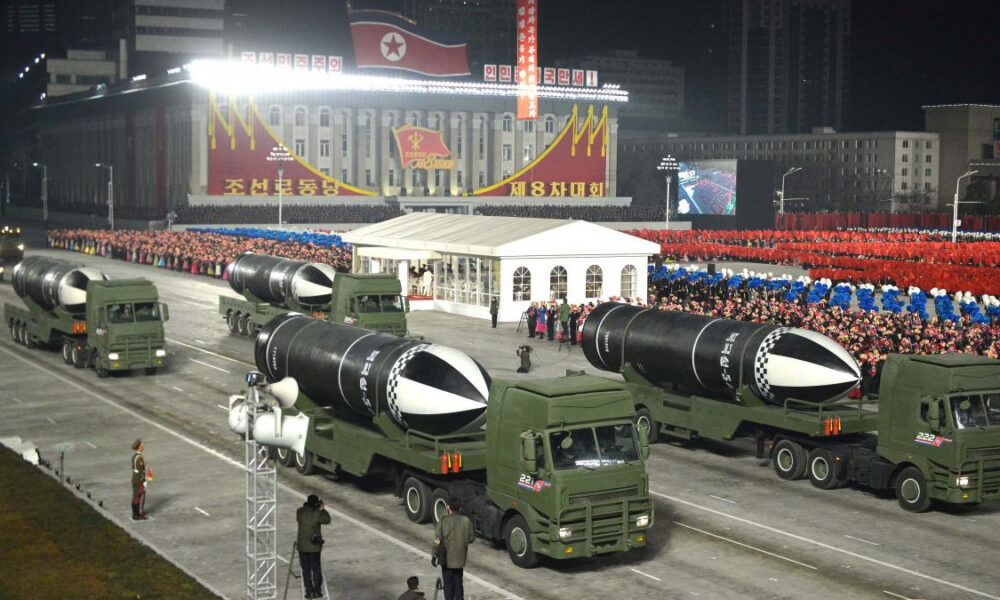 Night-time North Korean parade with several missile-carrying trucks passing by a large crowd and a government building in the background.