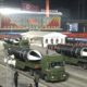 Night-time North Korean parade with several missile-carrying trucks passing by a large crowd and a government building in the background.