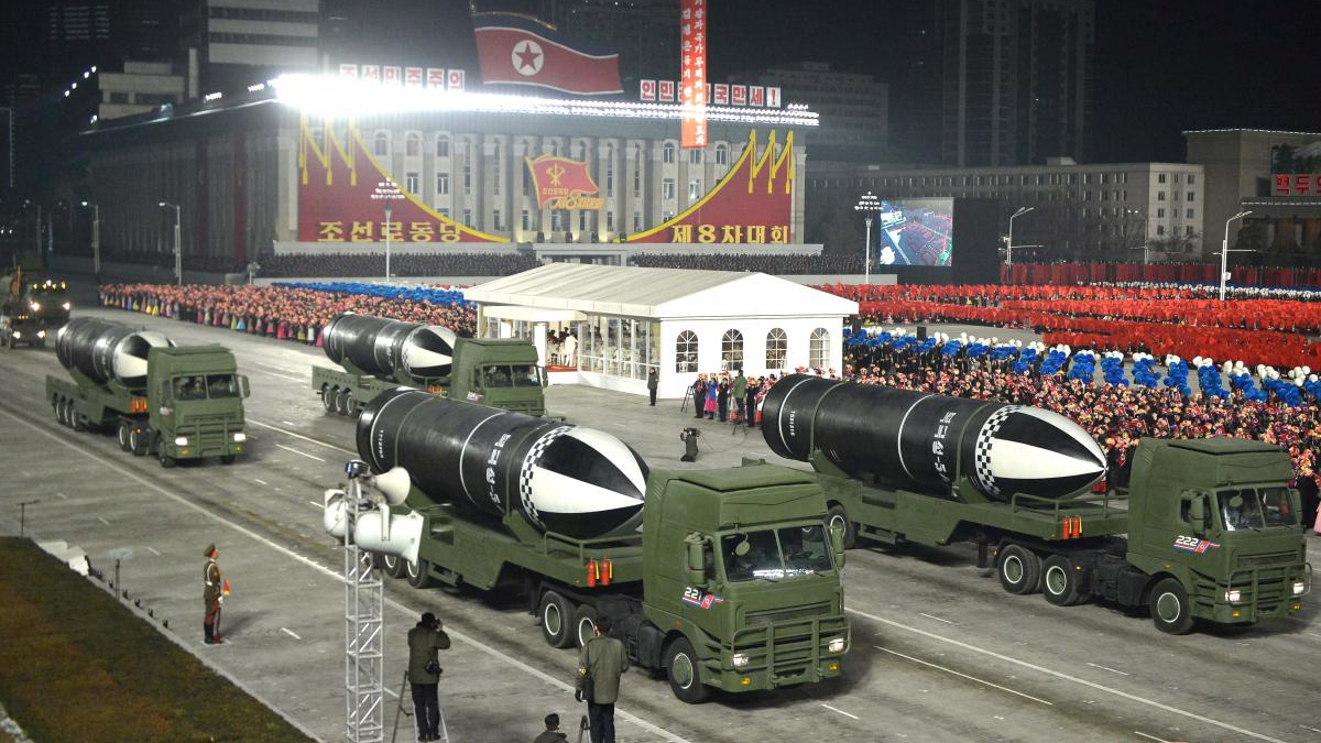 Night-time North Korean parade with several missile-carrying trucks passing by a large crowd and a government building in the background.