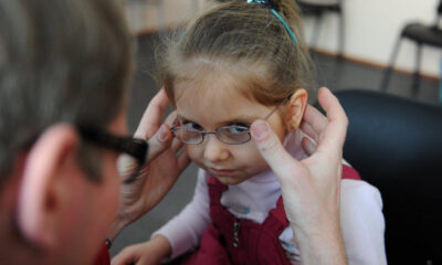 Adult helping a young girl fit her glasses, with both hands on the frame as the girl looks downward.