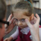 Adult helping a young girl fit her glasses, with both hands on the frame as the girl looks downward.