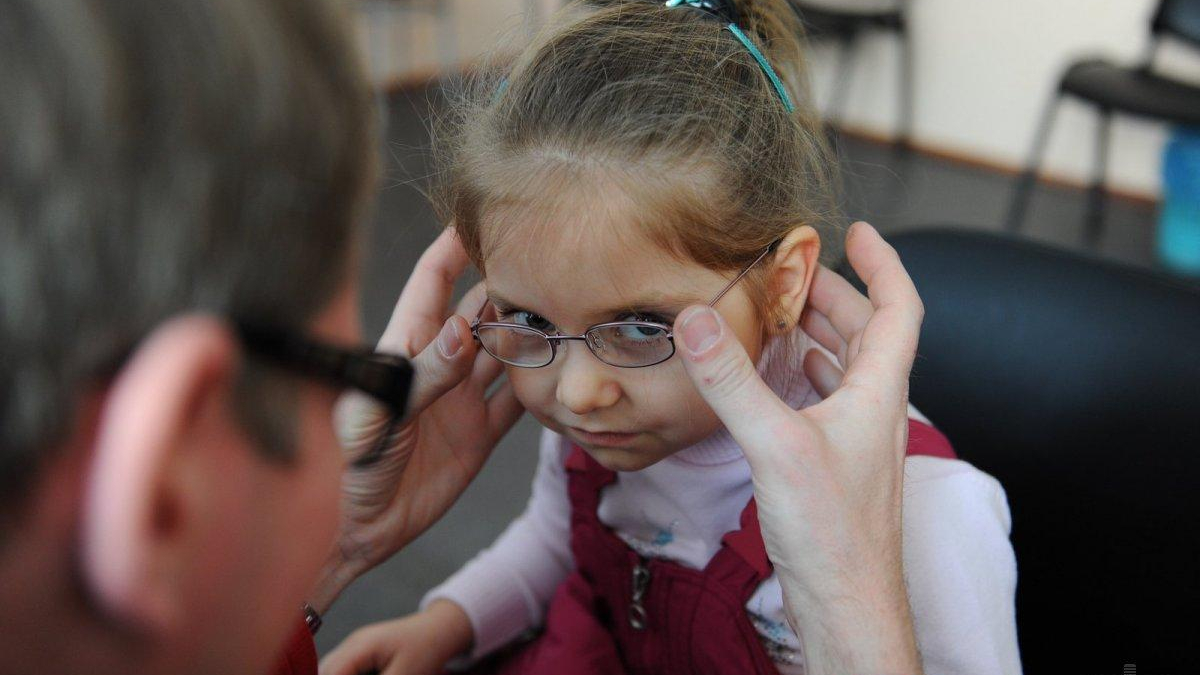Adult helping a young girl fit her glasses, with both hands on the frame as the girl looks downward.