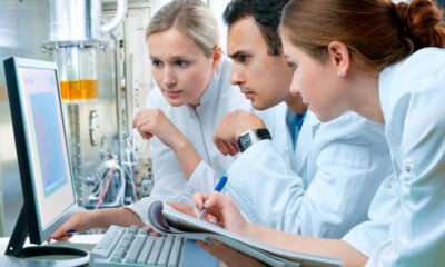 Three researchers in lab coats study data on a desktop computer in a high-tech laboratory setting.