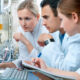 Three researchers in lab coats study data on a desktop computer in a high-tech laboratory setting.