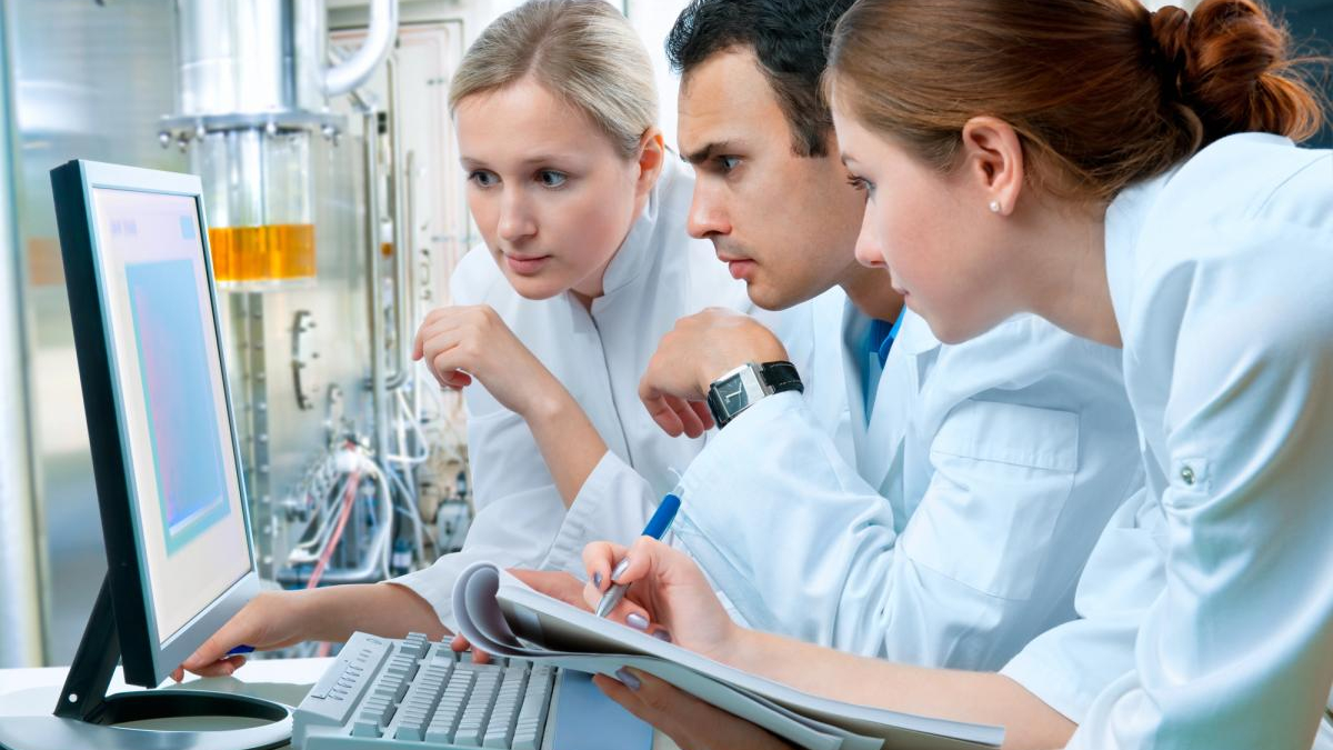 Three researchers in lab coats study data on a desktop computer in a high-tech laboratory setting.
