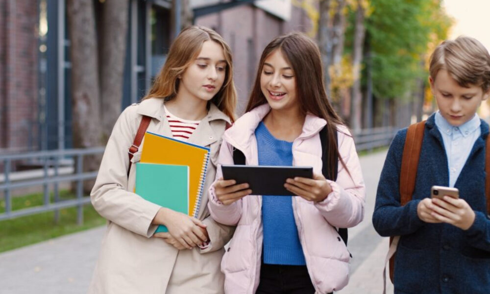 Three students walk together on a campus sidewalk; two girls carry notebooks while the third student looks at a tablet and a phone.