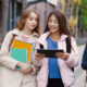 Three students walk together on a campus sidewalk; two girls carry notebooks while the third student looks at a tablet and a phone.