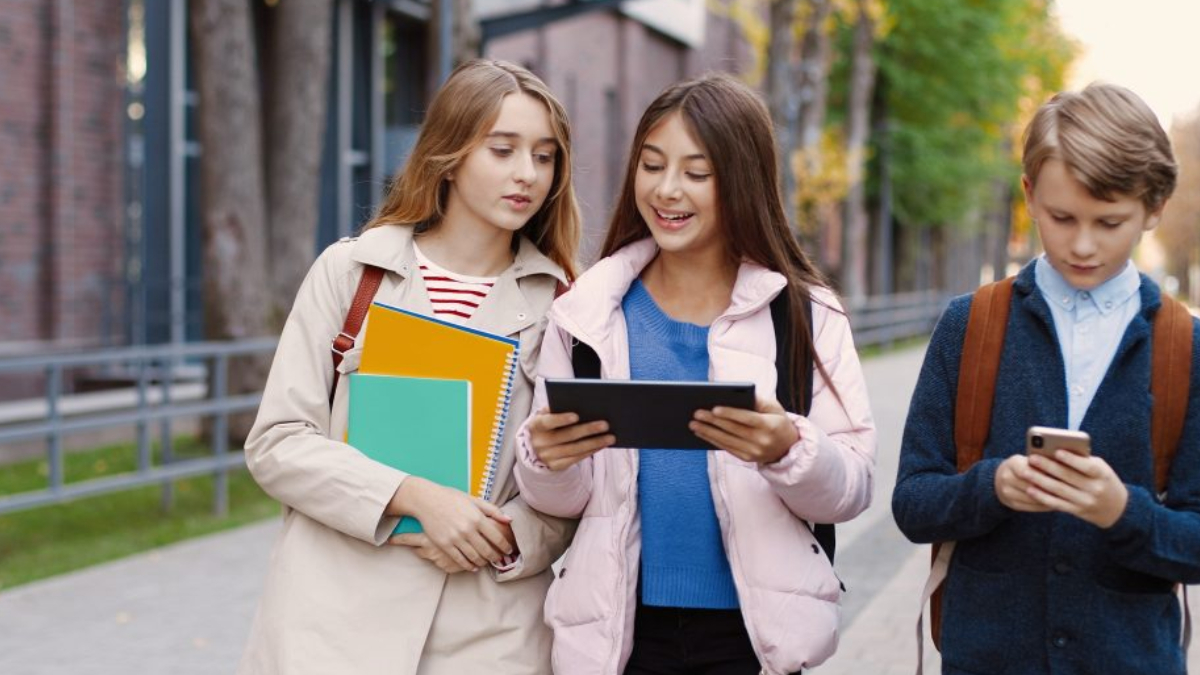 Three students walk together on a campus sidewalk; two girls carry notebooks while the third student looks at a tablet and a phone.