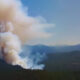 Large column of smoke rises from a forested valley with mountains in the distance under a blue sky.