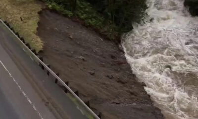 Aerial view of a road with a guardrail on the left, showing an eroded muddy riverbank next to turbulent water on the right.