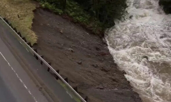 Aerial view of a road with a guardrail on the left, showing an eroded muddy riverbank next to turbulent water on the right.
