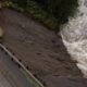 Aerial view of a road with a guardrail on the left, showing an eroded muddy riverbank next to turbulent water on the right.