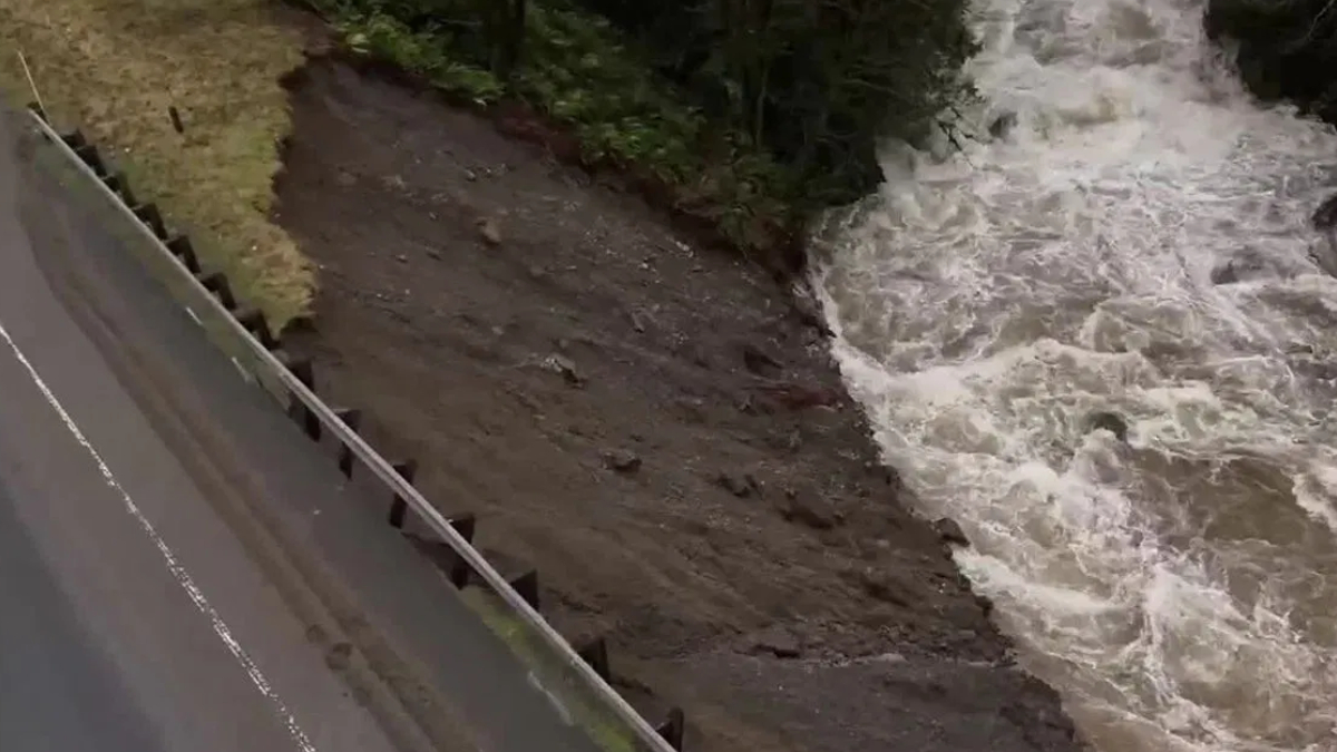Aerial view of a road with a guardrail on the left, showing an eroded muddy riverbank next to turbulent water on the right.