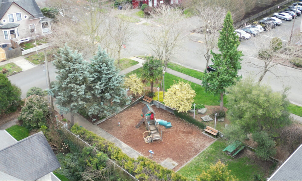 Aerial view of a small playground with a wooden structure and blue slide surrounded by mulch and benches under trees in a residential yard