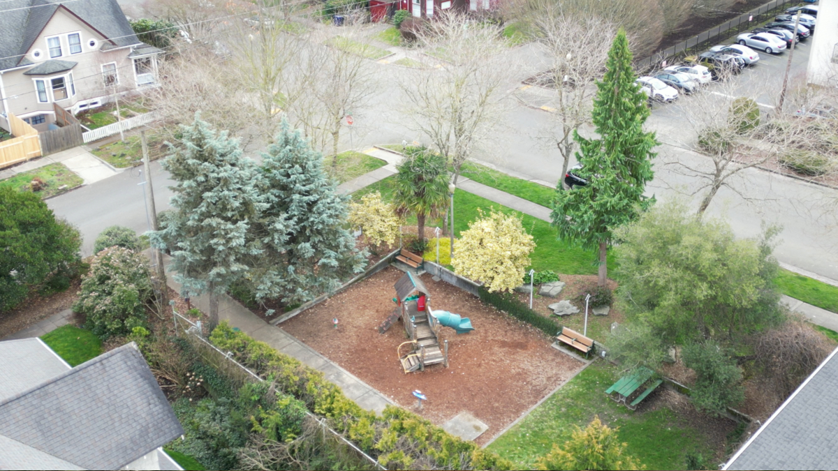 Aerial view of a small playground with a wooden structure and blue slide surrounded by mulch and benches under trees in a residential yard