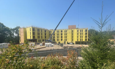Construction site with a multi-story building wrapped in yellow sheathing and a crane overhead lifting materials under a clear blue sky