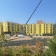 Construction site with a multi-story building wrapped in yellow sheathing and a crane overhead lifting materials under a clear blue sky