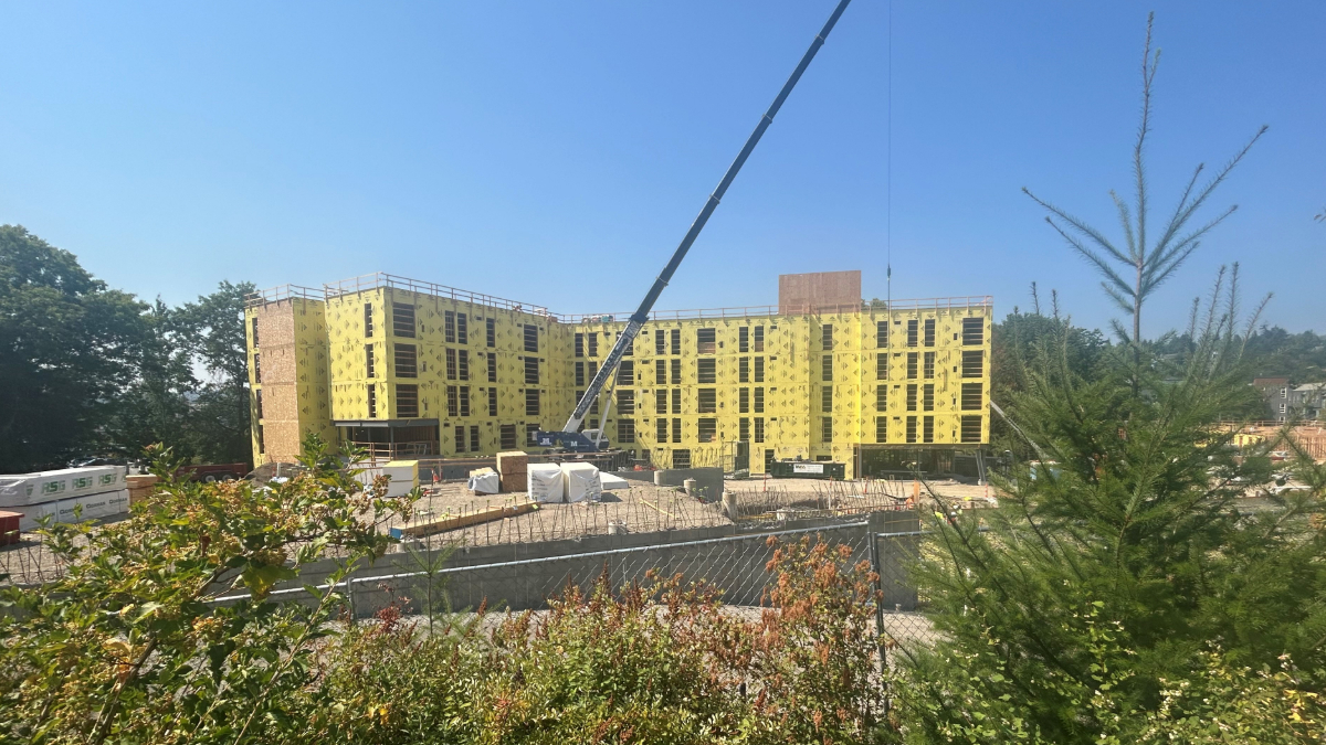 Construction site with a multi-story building wrapped in yellow sheathing and a crane overhead lifting materials under a clear blue sky
