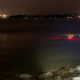 Nighttime waterfront: a swimmer in dark water near rocks and a wooden railing, city lights across the bay and a bridge to the right.