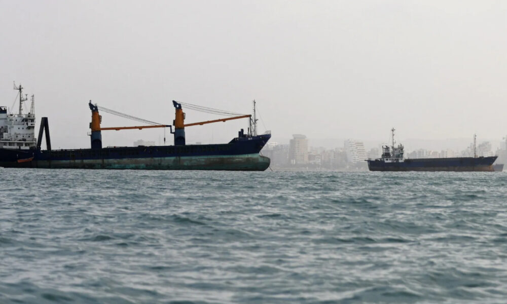 Large cargo ship with two orange cranes sailing near a city skyline, calm sea in foreground.