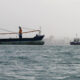 Large cargo ship with two orange cranes sailing near a city skyline, calm sea in foreground.