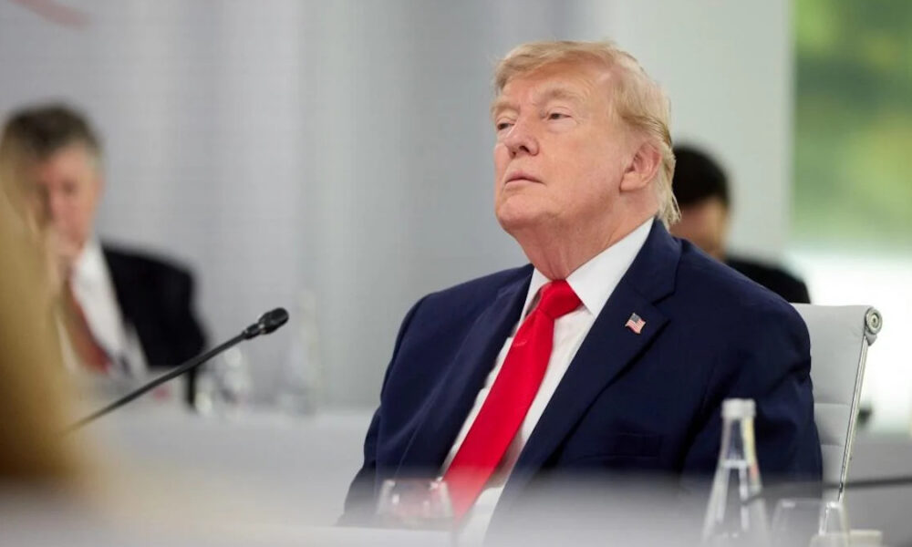 Former US president Donald Trump sits at a conference table in a navy suit and red tie, looking upward with a microphone in front of him.