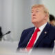 Former US president Donald Trump sits at a conference table in a navy suit and red tie, looking upward with a microphone in front of him.