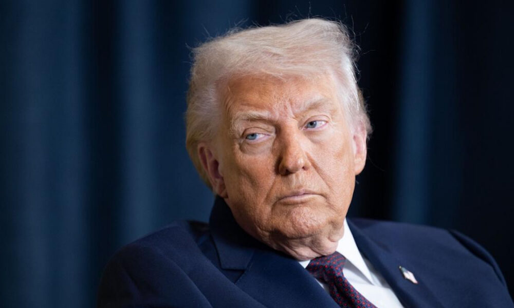 Older man in a dark navy suit and red tie looks to the left with a serious expression against a dark backdrop.