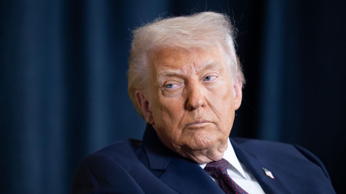 Older man in a dark navy suit and red tie looks to the left with a serious expression against a dark backdrop.