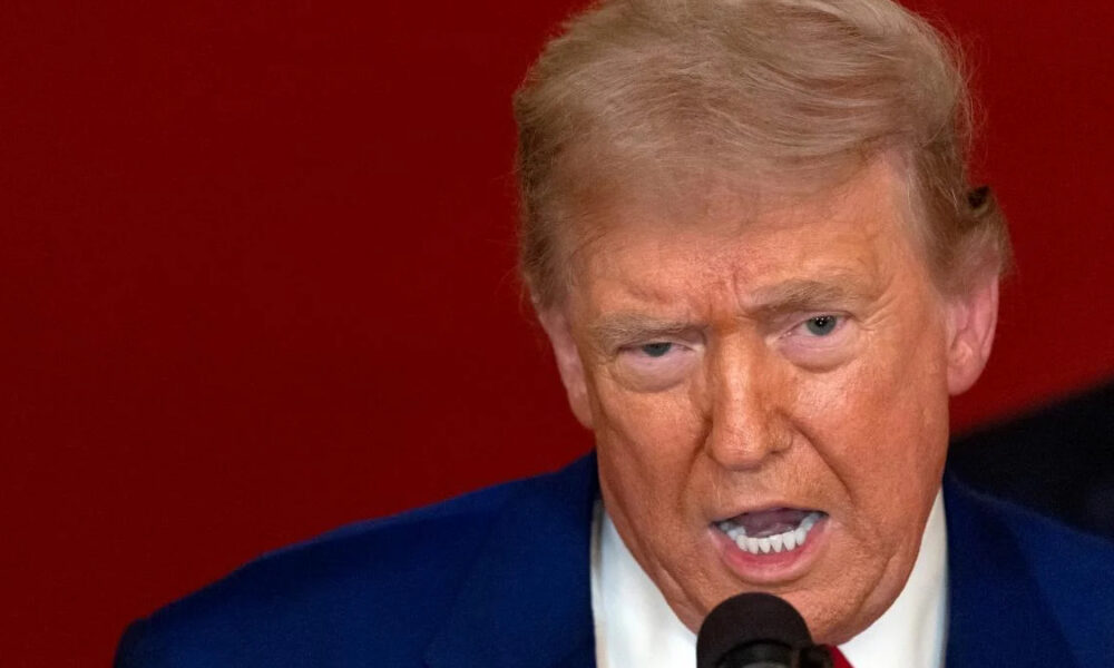 Close-up of a man in a blue suit speaking into a microphone against a red background, with a tense expression.
