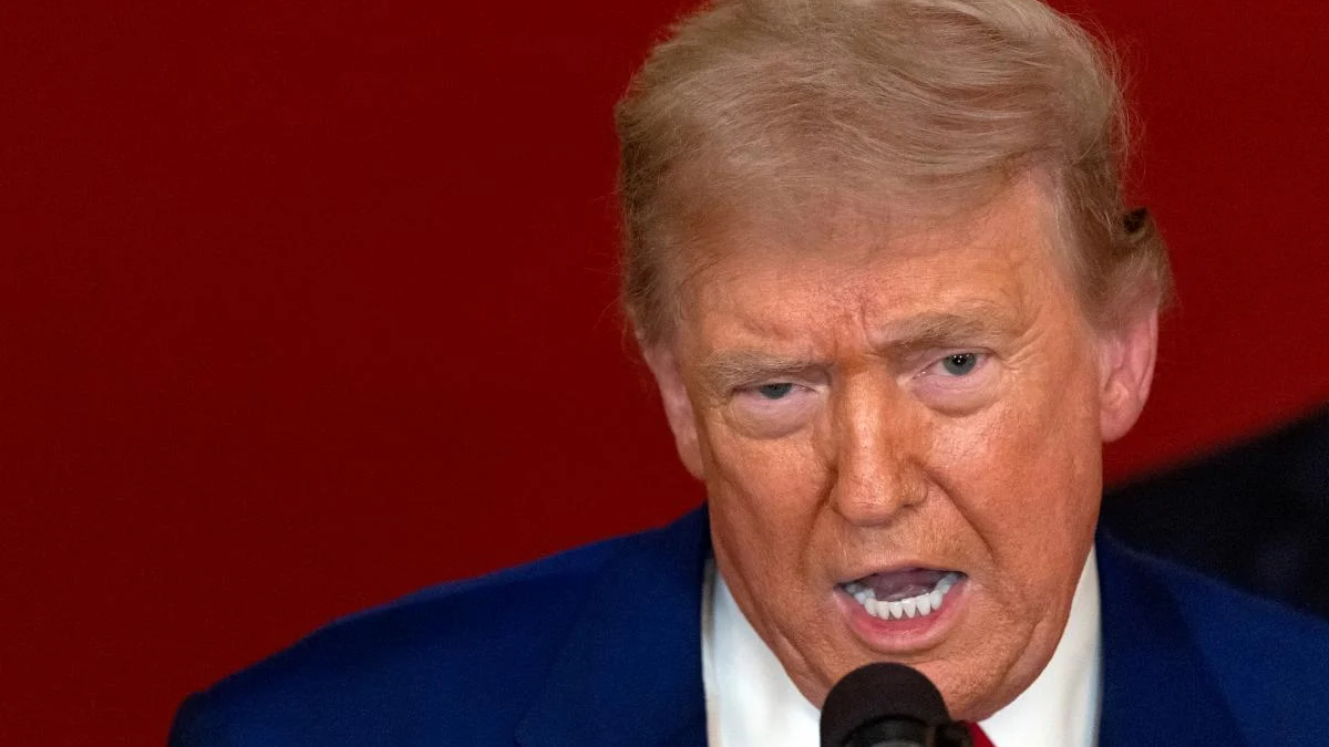 Close-up of a man in a blue suit speaking into a microphone against a red background, with a tense expression.