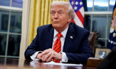 Former U.S. president Donald Trump seated at a desk with hands folded, wearing a navy suit and red tie, American flag behind him.