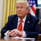 Former U.S. president Donald Trump seated at a desk with hands folded, wearing a navy suit and red tie, American flag behind him.