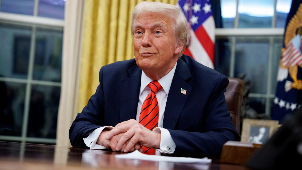 Former U.S. president Donald Trump seated at a desk with hands folded, wearing a navy suit and red tie, American flag behind him.