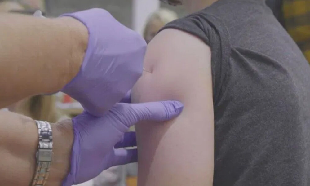 Healthcare worker with purple gloves giving a vaccine shot to a person's upper arm in a clinic setting.