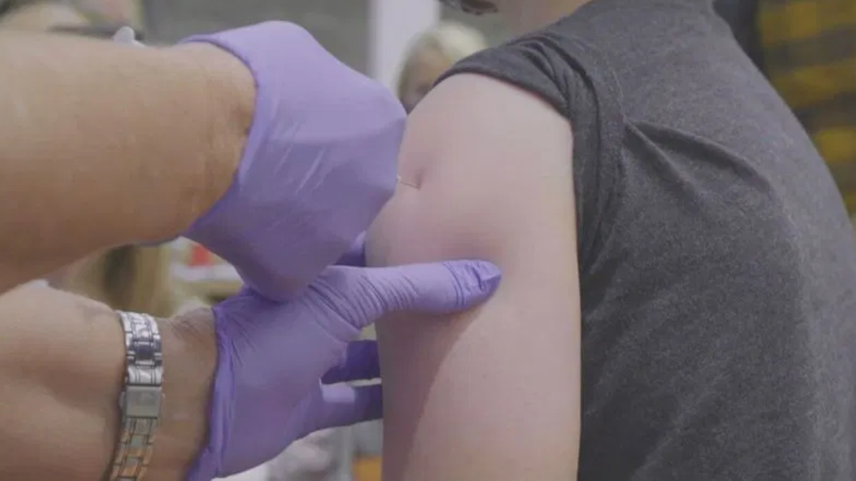 Healthcare worker with purple gloves giving a vaccine shot to a person's upper arm in a clinic setting.