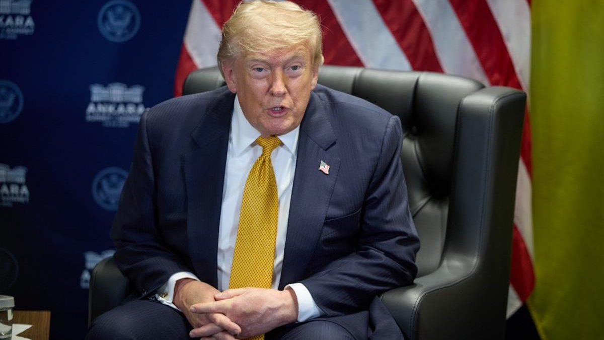 Man in a navy suit and yellow tie sits in a black leather chair, speaking, with American flags in the background.
