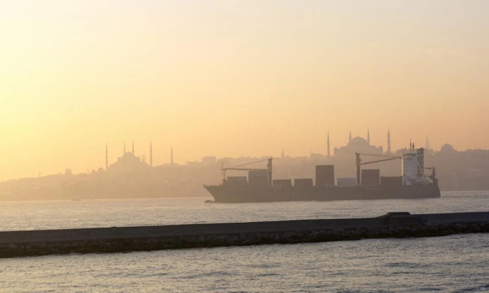 Container ship sailing at sunset on calm water with a distant, hazy city skyline.