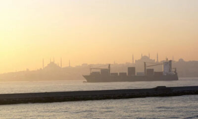 Container ship sailing at sunset on calm water with a distant, hazy city skyline.
