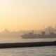 Container ship sailing at sunset on calm water with a distant, hazy city skyline.