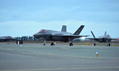 Two gray F-22 fighter jets parked on a runway at an airbase under a blue cloudy sky.