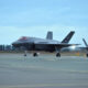 Two gray F-22 fighter jets parked on a runway at an airbase under a blue cloudy sky.
