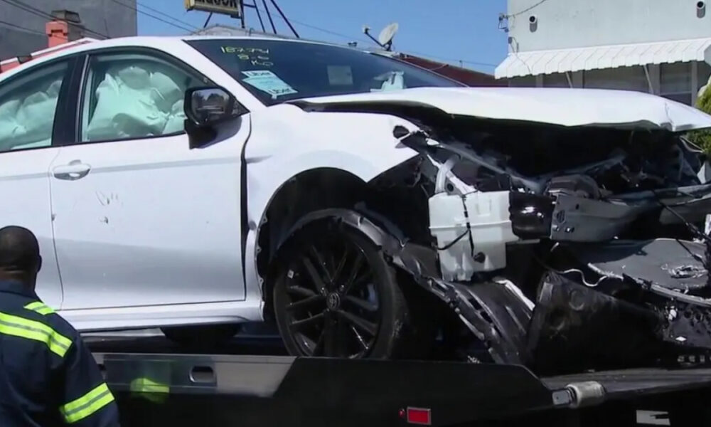 White sedan with severe front-end damage and deployed airbags on a tow truck bed, emergency responder nearby in a neon jacket.