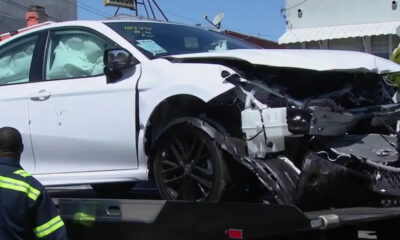 White sedan with severe front-end damage and deployed airbags on a tow truck bed, emergency responder nearby in a neon jacket.